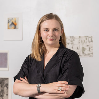 Woman in a black shirt standing with arms crossed against a white wall with framed pictures.