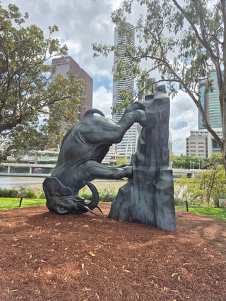 Statue of an elephant on a ruck, turned to 90 degrees so that it's head is against the ground, in a park with city skyline in the background.